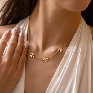 May include: A gold-toned chain necklace featuring three evenly spaced paw print charms. Each charm is engraved with a name, including Leo, Milo, and Bella. The delicate metal chain connects the charms in a horizontal row across the collarbone area.