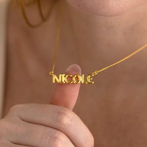 Könnte beinhalten: Eine goldfarbene Halskette mit dem Namen "NICOLE" in Großbuchstaben. Der Anhänger hängt an einer feinen Kette. Das Bild ist eine Nahaufnahme, die das Design der Halskette und die Bedeutung des Namens hervorhebt.