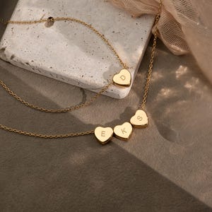 Könnte beinhalten: Zwei goldfarbene Halsketten mit herzförmigen Anhängern. Eine Kette hat einen einzelnen Anhänger mit dem eingravierten Buchstaben D. Die zweite Kette hat drei herzförmige Anhänger mit den Buchstaben E, K und S. Beide Ketten haben feine Glieder.