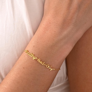 May include: A gold-toned metal bracelet featuring the words "Healthy food Only" written in a cursive script font. The text is connected by small metal links, forming a continuous chain that wraps around the wrist with a delicate, thin design.