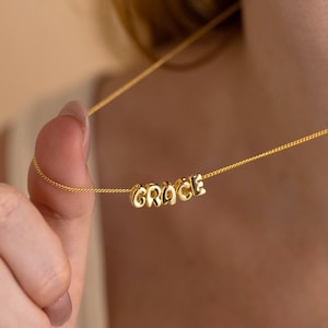 May include: A gold-colored necklace with the name "GRACE" spelled out in individual, rounded letters. The necklace is on a delicate chain. The image is a close-up, showing the necklace against a blurred background.