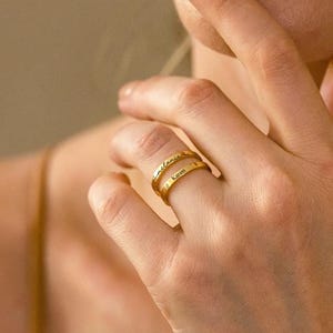 May include: Two gold rings, one stacked on top of the other, are shown on a finger. The top ring has the name "melania" engraved on it, and the bottom ring has the name "Karen" engraved on it. The rings are displayed against a soft, neutral background.