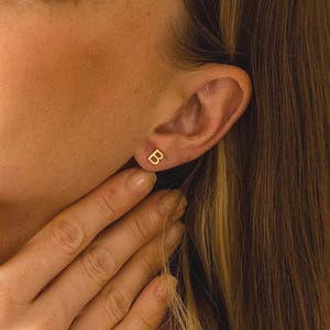 Könnte beinhalten: Eine Frau mit langen blonden Haaren lächelt und trägt kleine goldene quadratische Ohrstecker. Die Ohrringe sind in ihrem Ohr sichtbar. Sie berührt ihren Hals mit ihrer Hand. Der Hintergrund ist einfarbig beige.