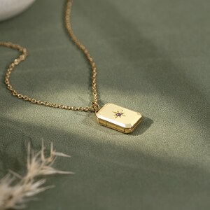 May include: A gold-toned rectangular locket pendant suspended from a delicate matching chain. The locket features a polished surface with a central starburst engraving, accented by a small, embedded purple gemstone at its very center.
