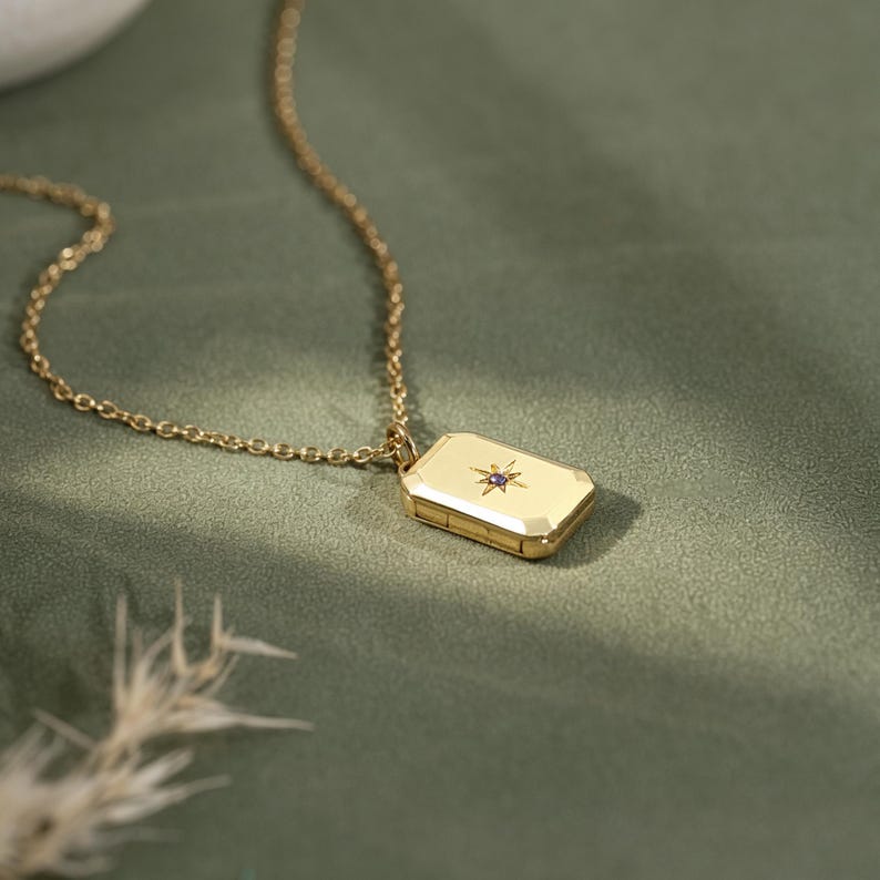 May include: A gold-toned rectangular locket pendant suspended from a delicate matching chain. The locket features a polished surface with a central starburst engraving, accented by a small, embedded purple gemstone at its very center.