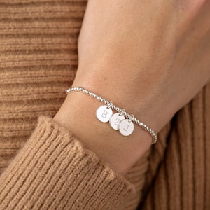 May include: Silver beaded bracelet featuring three circular charms, each engraved with a single initial. The bracelet is displayed on a wrist, against a backdrop of a brown jumper.