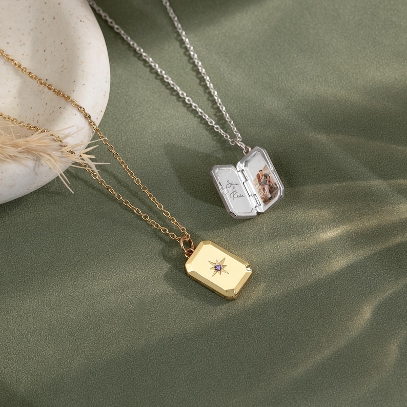 May include: Two rectangular locket necklaces with thin chains. One gold-toned locket features a starburst engraving with a small purple stone. The other silver-toned locket is open, revealing a photograph of two people and cursive text inside.