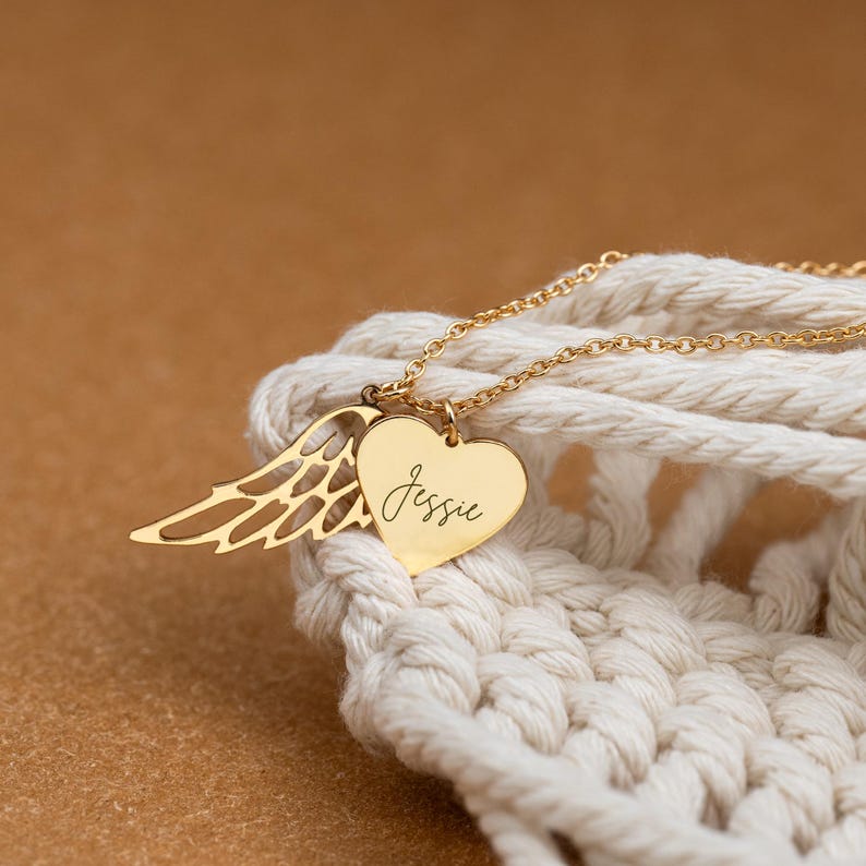 May include: A gold-colored necklace with a heart-shaped pendant and a wing charm. The heart pendant is inscribed with the name "Jessie" in cursive script. The necklace is displayed on a white, textured surface.