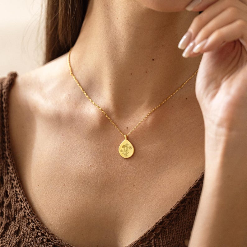 Op de afbeelding: Een goudkleurige ketting met een druppelvormige hanger met een bloemmotief. De hanger hangt aan een fijne ketting. De ketting wordt gedragen over een bruine top.