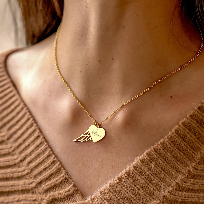 May include: A gold-colored necklace with a delicate chain. The pendant features a heart and a wing charm. The heart charm has the name "Monica" engraved on it. The necklace is worn against a brown sweater.