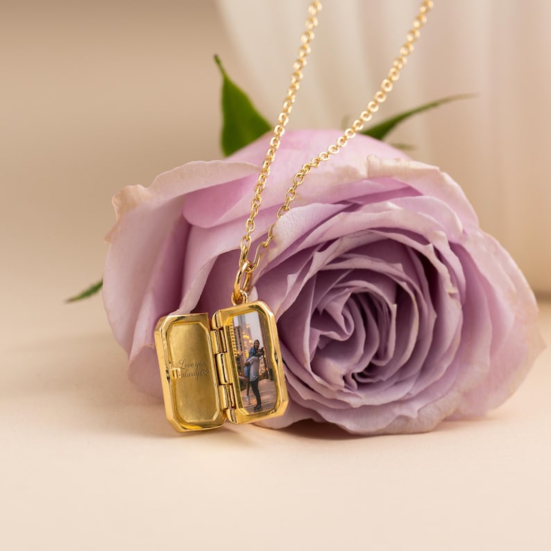 May include: A gold-toned metal locket necklace with a delicate chain. The rectangular locket is open, revealing a small photograph of two people on the right side and the engraved text 'Love you always' on the left interior panel of the locket.