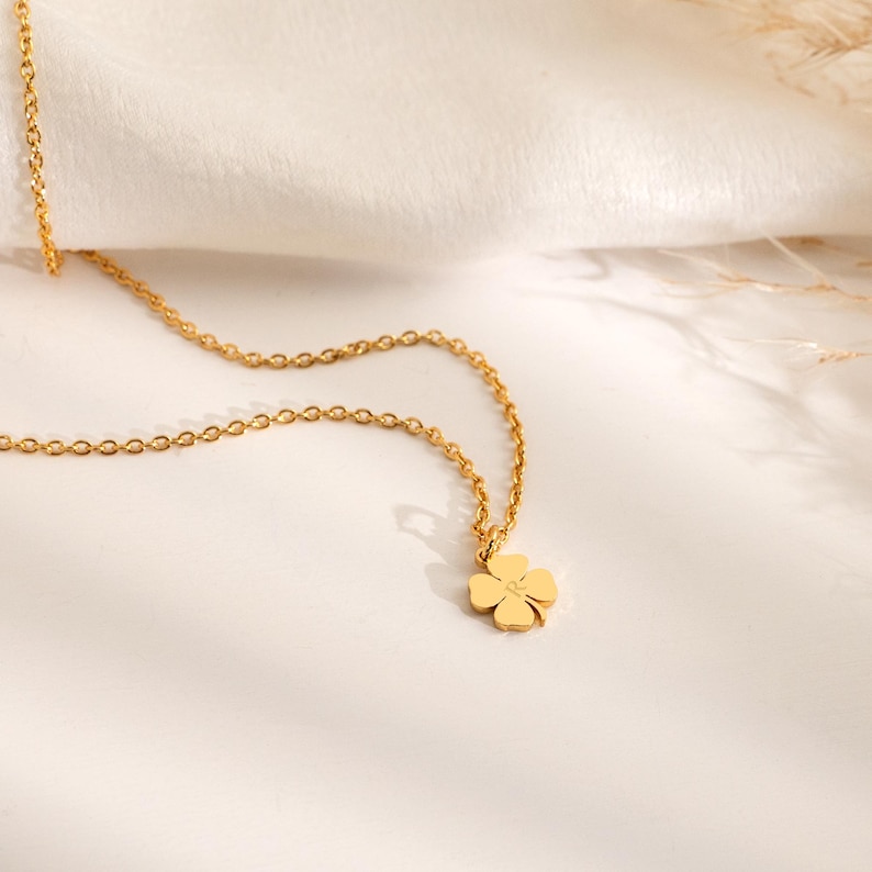 May include: A gold-colored necklace with a four-leaf clover pendant. The chain is delicate and the clover has a small, centered detail. The necklace is displayed on a white surface with soft lighting.