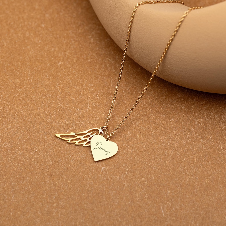 May include: A gold-colored necklace with a heart pendant and a wing charm. The heart pendant has the name "Dennis" engraved on it. The necklace is displayed on a neutral-toned background.