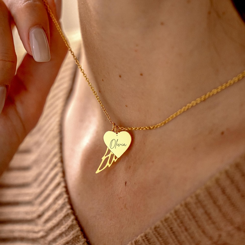 May include: A gold-colored necklace featuring a heart-shaped pendant with the name "Olivia" inscribed on it. The pendant is paired with a wing-shaped charm. The necklace is displayed on a person's neck.