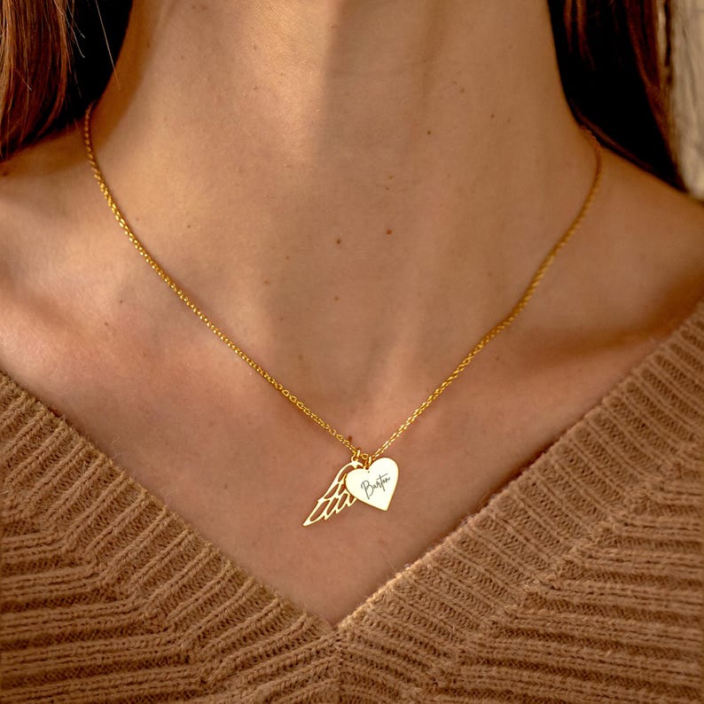 May include: Gold-colored necklace with a heart pendant and a wing charm. The heart pendant has the word "Bestie" written on it. The necklace is worn on a person wearing a brown sweater.