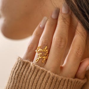 Può includere: Un anello color oro caratterizzato da due righe sovrapposte di testo in corsivo. La riga superiore mostra il nome Emily e la riga inferiore mostra il nome Jayde. La fascia metallica è sottile e segue il contorno delle lettere in corsivo.