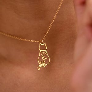 May include: A gold-colored necklace with a cat pendant. The pendant is a silhouette of a cat with the name "Riley" inscribed in a cursive font. The necklace is worn around a person's neck.