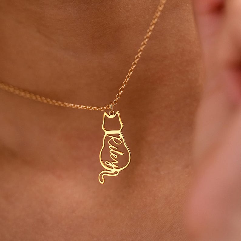 May include: A gold-colored necklace with a cat pendant. The pendant is a silhouette of a cat with the name "Riley" inscribed in a cursive font. The necklace is worn around a person's neck.