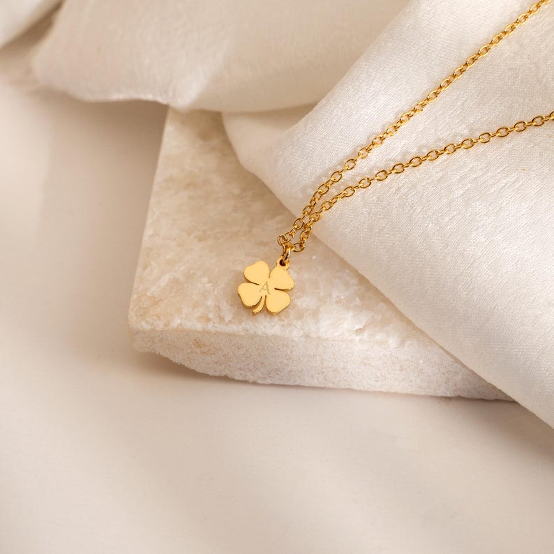 May include: A gold-colored necklace with a four-leaf clover pendant. The clover is centered and hangs from a delicate chain. The necklace is displayed on a white surface with a soft, draped fabric in the background. The clover pendant has the letter "A" in the center.