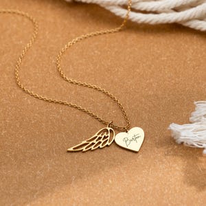 May include: A gold-colored necklace with a delicate chain. The necklace features a wing charm and a heart-shaped pendant with the name "Barton" engraved on it. The background is a warm, neutral tone.