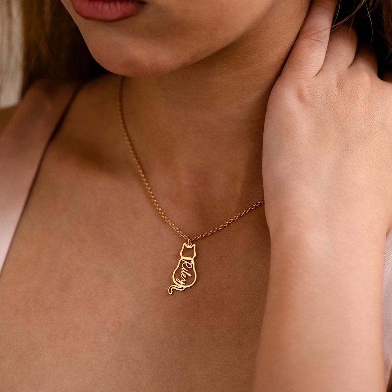May include: A gold-colored necklace featuring a cat pendant. The pendant has the name "Riley" inscribed on it. The necklace is worn around the neck. The background is a soft, neutral color.