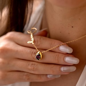 May include: A gold necklace featuring a teardrop-shaped, dark purple gemstone pendant. The necklace has a delicate chain and a cursive name charm. The pendant is set in a gold bezel. The necklace is being held up.