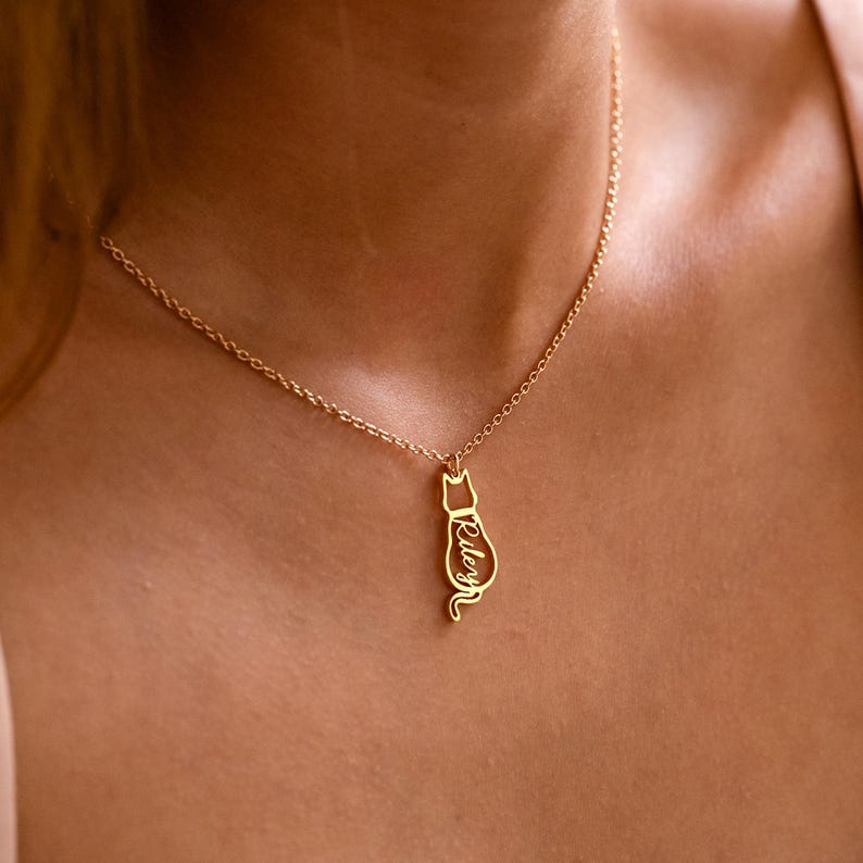 May include: A gold-colored necklace featuring a cat pendant. The pendant is a silhouette of a cat with the name "Riley" inscribed on it. The necklace is worn around the neck, showcasing the delicate chain and pendant.
