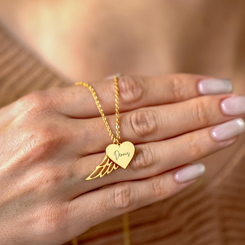 May include: A gold-colored necklace with a heart-shaped pendant and a wing design. The heart pendant has the name "Dennis" engraved on it. The necklace is held in a hand.