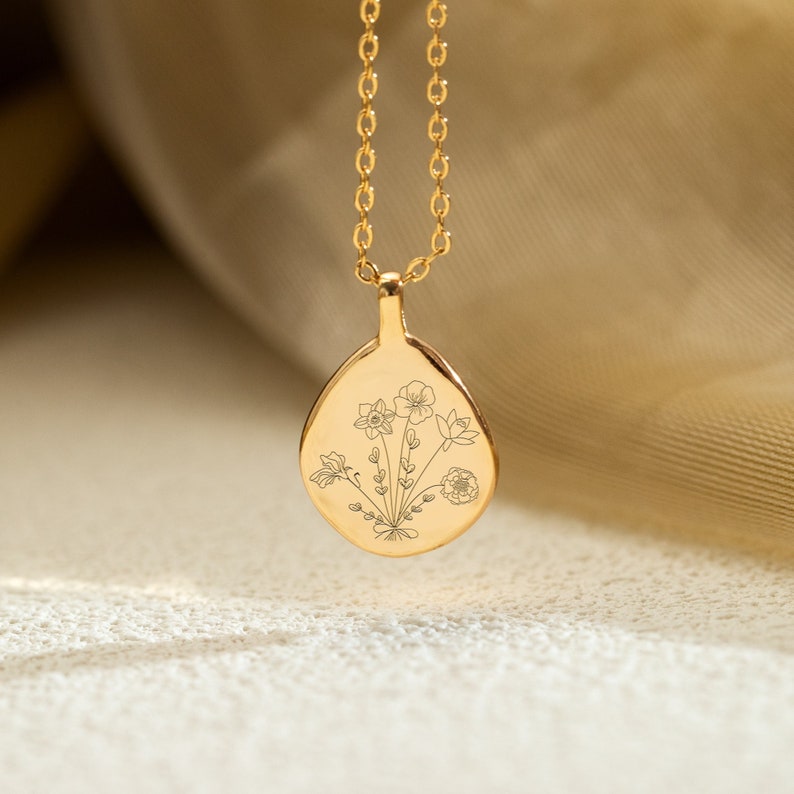 Op de afbeelding: Een goudkleurige hangerketting met een druppelvormige bedel. De bedel heeft een gegraveerd bloemenontwerp. De ketting en hanger zijn gemaakt van glanzend, goudkleurig metaal. De achtergrond is een zachte, neutrale kleur.