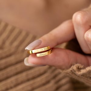 May include: A gold-coloured wrap ring engraved with the names "Olivia" and "Ethan". The ring is displayed on a finger, with the names clearly visible. The ring has a simple, elegant design.