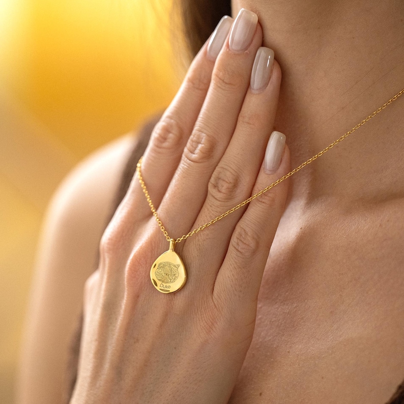 K&ouml;nnte beinhalten: Eine goldfarbene Halskette mit einem tropfenf&ouml;rmigen Anh&auml;nger. Der Anh&auml;nger hat ein detailliertes Design und das Wort "Oaks" unten. Die Halskette wird um den Hals getragen, wobei die Kette sichtbar ist. Der Hintergrund ist ein weiches Gelb.