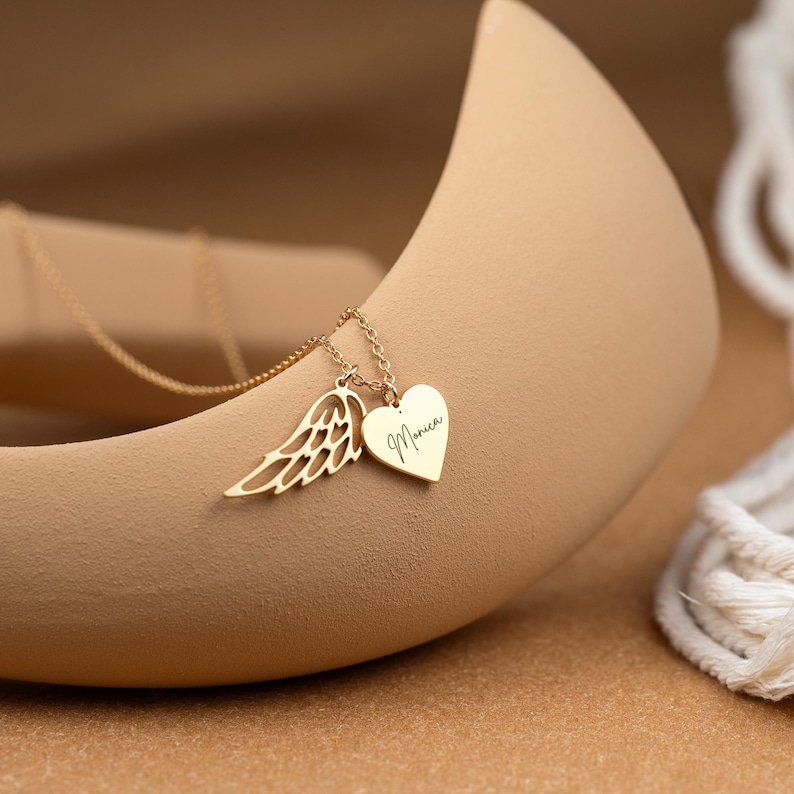 May include: A gold-colored necklace with a delicate chain. The pendant features a wing charm and a heart-shaped charm with the name "Monica" engraved. The necklace is displayed on a neutral-toned background.