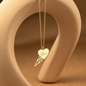 May include: A gold-colored necklace with a heart-shaped pendant and a wing design. The pendant has the name "Olivia" engraved on it. The necklace is hanging in front of a beige, abstract sculpture.