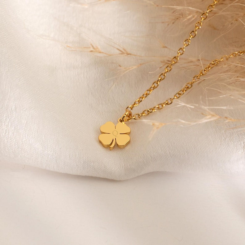 May include: A gold-colored necklace with a four-leaf clover pendant. The chain is delicate, and the clover is centered. The necklace is displayed on a white, textured surface with dried plants in the background. The jewelry is simple and elegant.
