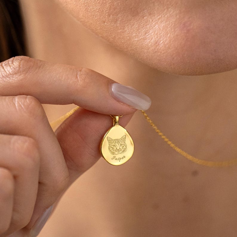 K&ouml;nnte beinhalten: Goldfarbene Halskette mit einem tropfenf&ouml;rmigen Anh&auml;nger. Der Anh&auml;nger zeigt ein eingraviertes Katzen-Gesicht und den Namen "Jasper" darunter. Die Halskette wird von einer Person gehalten.