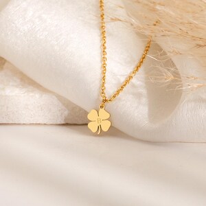 May include: A gold-colored necklace with a four-leaf clover pendant. The pendant has the letter "B" engraved in the center. The necklace is displayed on a white, textured surface.