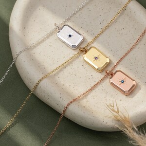 May include: Three rectangular pendant necklaces in silver, gold, and rose gold tones. Each pendant features a central starburst engraving with a small embedded gemstone: a dark blue stone in the silver, a purple stone in the gold, and a light blue stone in the rose gold.
