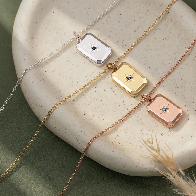 May include: Three rectangular pendant necklaces in silver, gold, and rose gold tones. Each pendant features a central starburst engraving with a small embedded gemstone: a dark blue stone in the silver, a purple stone in the gold, and a light blue stone in the rose gold.