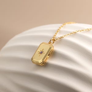 May include: A gold-toned rectangular locket pendant on a matching delicate chain. The locket features a beveled edge and a central starburst engraving with a small, embedded purple gemstone at its core, creating a polished metallic finish.