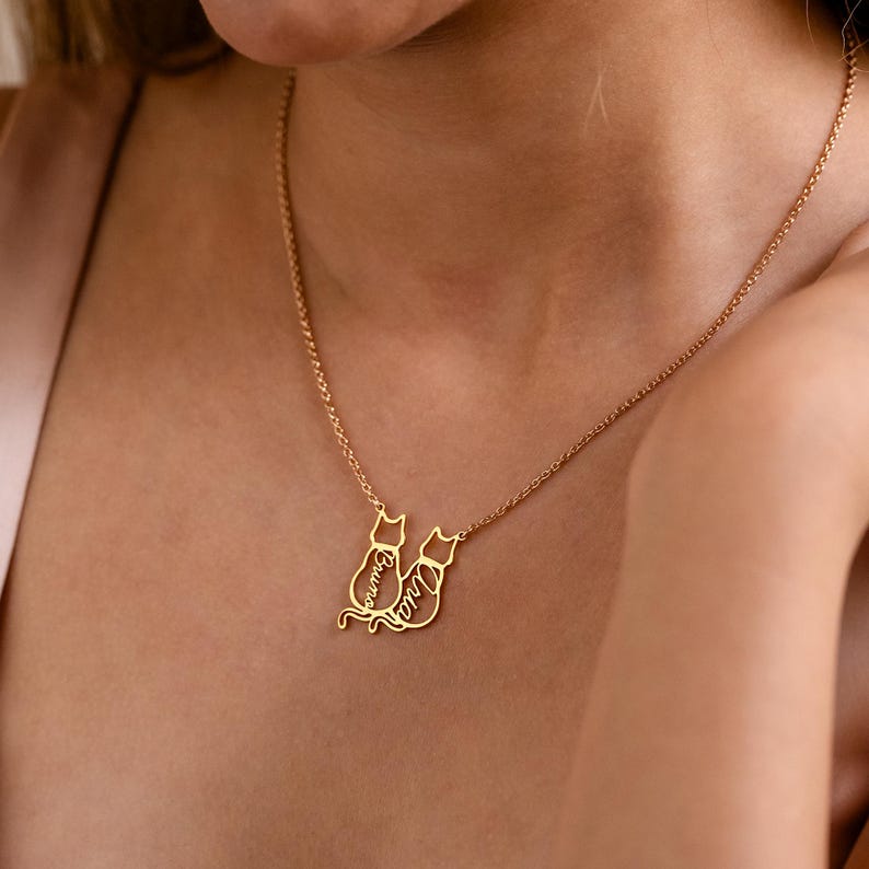May include: A gold-colored necklace featuring two cat silhouettes. Each cat has a name engraved on it. The necklace is worn around the neck and is a delicate piece of jewelry.