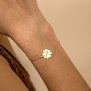 May include: Gold-colored bracelet featuring a four-leaf clover charm. The clover has four heart-shaped leaves, each with a small inscription. The bracelet is delicate and has a chain-link design.