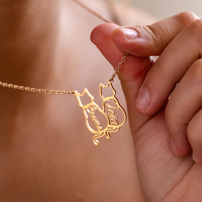 May include: Gold-colored necklace featuring two cat silhouettes. Each cat silhouette has a name engraved on it. The necklace is held by a person. The names on the cats are "Bruno" and "Aria".