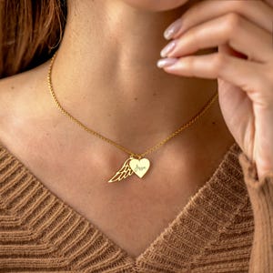 May include: A gold-colored necklace with a heart pendant and a wing charm. The heart pendant has the name "Jessie" engraved on it. The necklace is worn on a person with a brown sweater.