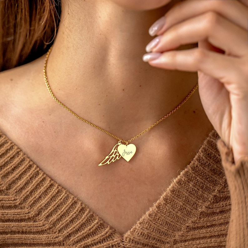May include: A gold-colored necklace with a heart pendant and a wing charm. The heart pendant has the name "Jessie" engraved on it. The necklace is worn on a person with a brown sweater.