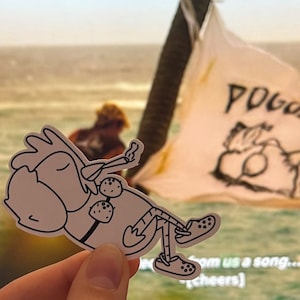 Puede incluir: Una pegatina blanca con un personaje de pollo de dibujos animados con Crocs y relajándose. La pegatina se sostiene frente a un fondo borroso de una escena de playa y una bandera con el texto "POGUELAND".