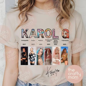 May include: A beige t-shirt featuring the name "KAROL G" in a colorful, block letter design. Below the name are images of Karol G with text indicating album names and years, including "Unstoppable," "Ocean," and "KG0516."
