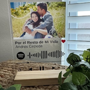 May include: Clear acrylic photo display featuring a couple embracing, with the text "Por el Resto de Mi Vida" and a Spotify-style music bar. The display is mounted on a wooden base, with a heart symbol. The photo is approximately 6 inches wide.