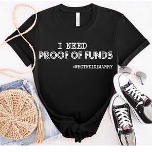 May include: Black t-shirt with white text that reads "I need proof of funds #WHOTFDIDIMARRY". The shirt is laid out on a white surface with a pair of black sneakers, a straw bag, and a pair of sunglasses.