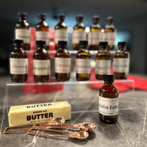 May include: A collection of small glass bottles with labels, arranged on a shelf. The bottles contain various extracts, including vanilla, almond, and chocolate. A stick of butter and measuring spoons are in the foreground.