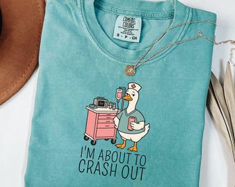 May include: A teal crewneck t-shirt featuring a graphic of a white duck dressed as a nurse, standing next to a pink medical cart with an IV drip and monitor. The duck holds a pink can, and the text below reads, I'M ABOUT TO CRASH OUT.